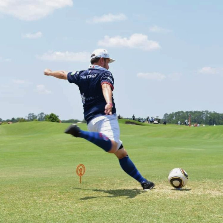 Joueur en uniforme bleu foncé et blanc donne un coup de pied à un ballon sur un terrain herbeux.