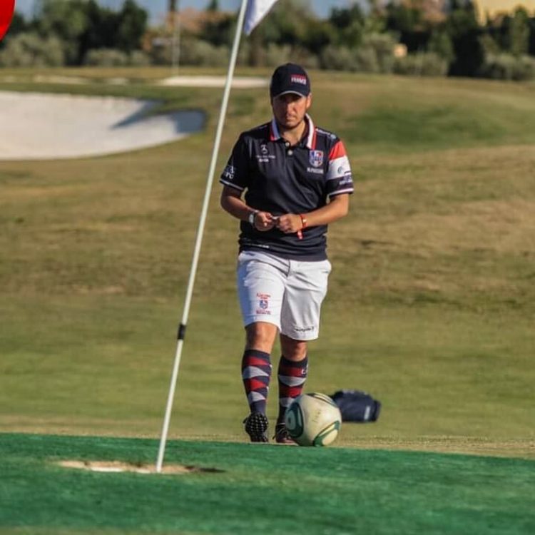 Homme en tenue sportive avec un ballon de football sur un green de golf près du drapeau.
