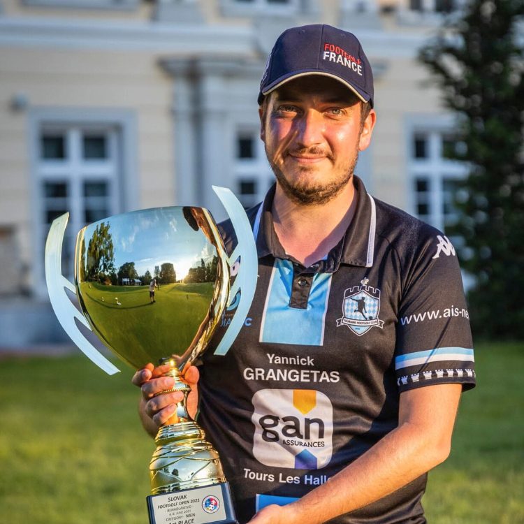 Homme en casquette tenant un trophée doré devant un bâtiment avec pelouse verte.